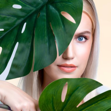 Closeup facial portrait personable woman with perfect smooth makeup holding green monstera leaves and cover her face as natural healthy skincare treatment. Tropical nature and beauty concept