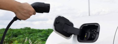 Progressive natural scenic with windmill generator where hand insert charging plug to electric vehicle from charging station with natural background. EV car powered by wind turbine electric generator.