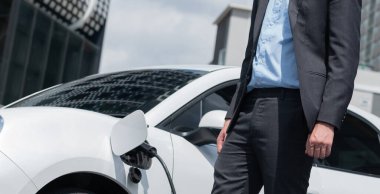 Closeup progressive businessman with electric car recharging at public charging station with background of city residential building. Eco friendly car powered by alternative clean energy.