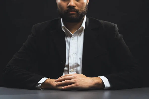 Businessman or lawyer wearing formal black suit sitting at table on isolated black background. Concept of a man with authority and seriousness gesture. equility