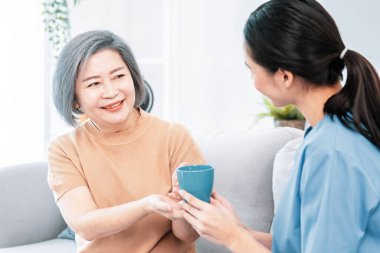 Female care taker serving her contented senior patient with a cup of coffee at home, smiling to each other. Medical care for pensioners, Home health care service.