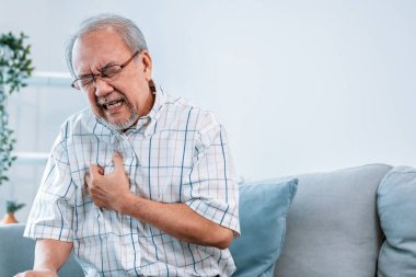 An agonizing senior man suffering from chest pain or heart attack alone in his living room. Serious health problem and feeling unwell concept for seniors.