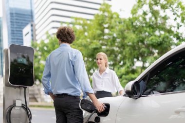 Progressive businessman and businesswoman with electric car parking and connected to public charging station before driving around city center. Eco friendly rechargeable car powered by clean energy.