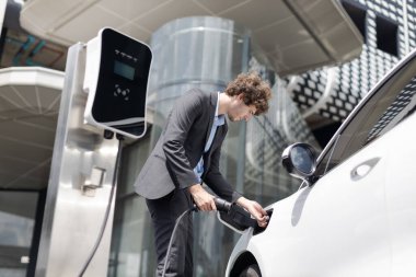 Below view closeup image of progressive black suit businessman recharge battery of his electric vehicle from public charging station. Renewable and alternative energy powered car concept.
