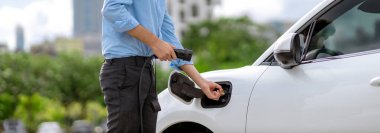 Closeup progressive man holding EV charger plug from public charging station for electric vehicle with background of residential building as concept eco-friendly sustainability energy car concept.