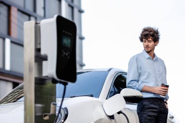 Progressive eco-friendly concept of parking EV car at public electric-powered charging station in city with blur background of businessman leaning on recharging-electric vehicle with coffee.