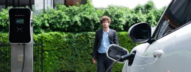 Progressive businessman wearing black suit with electric car recharging at public parking car charging station at modern city residential area. Eco friendly rechargeable car powered by clean energy.