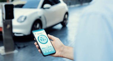 Closeup progressive businessman look at EV cars battery status application on smart phone screen at public parking charging station with power cable plug and renewable energy-powered electric vehicle