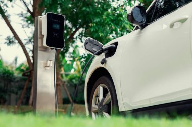 Focus closeup electric vehicle plugged in with EV charger device from blurred background of public charging station powered by renewable clean energy for progressive eco-friendly car concept.