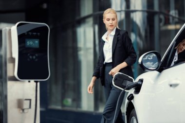 Arka planda ilerleyen iş kadınının yürüdüğü elektrikli araba için kamu EV şarj istasyonuna odaklan. Ekolojik kaygılar için temiz ve sürdürülebilir enerjiyle çalışan elektrikli araba..