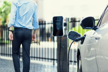 Closeup progressive suit-clad businessman with his electric vehicle recharge his car on public charging station in modern city with power cable plug and renewable energy-powered electric vehicle.
