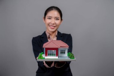 Resmi takım elbise giyen genç Asyalı kadın izole edilmiş arka planda ev modelini tutuyor ve sergiliyor. Konut ilanı, ev sigortası promosyonları ya da kişiselleştirilebilir fotokopi alanı için ideal. Coşkulu.