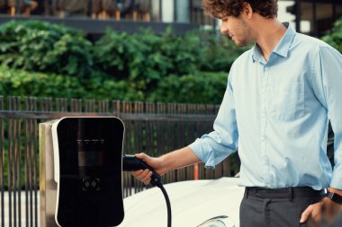 Progressive businessman insert charger plug from charging station to his electric vehicle with apartment condo building in background. Eco friendly rechargeable car powered by sustainable energy.