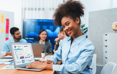 Bir grup ofis çalışanı ile birlikte arka planda ekranda gösterim paneliyle buluşan mutlu Afrikalı genç bir iş kadını portresi. Takım toplantısında kendine güveni tam bir ofis hanımı. Uyum