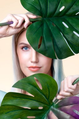 Closeup facial portrait personable woman with perfect smooth makeup holding green monstera leaves and cover her face as natural healthy skincare treatment. Tropical nature and beauty concept
