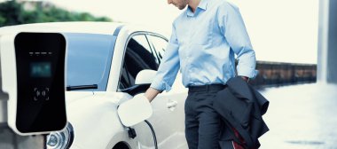 Closeup progressive man holding EV charger plug from public charging station for electric vehicle with background of residential building as concept eco-friendly sustainability energy car concept.