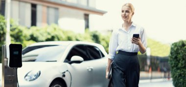 İş kadını tablet kullanıyor, halka açık otoparkta elektrikli aracını şarj ederken yürüyor. Teknolojinin ilerici yaşam tarzı ve EV otomobilinin ekolojik kaygısı