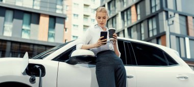 Focus businessman using phone, leaning on electric vehicle, holding coffee with blurred city residential condo buildings in background as progressive lifestyle by renewable and sustainable EV car.