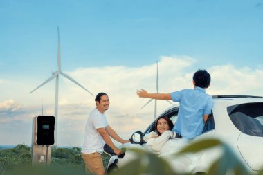 İlerici mutlu aile kavramı elektrikli araçla rüzgar çiftliğinde eğleniyor. Rüzgar türbini jeneratöründen gelen temiz yenilenebilir enerji ile şarj edilen elektrikli araç..