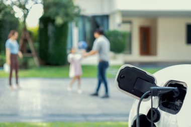 Focus EV charger connected with EV car at outdoor home charging station with blur progressive family playing together in the background. EV car for alternative clean energy concept.