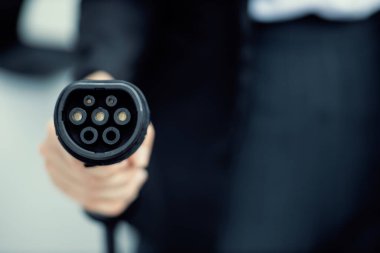 Focus closeup hand holding EV plug for electric vehicle pointing at camera with charging station at background as progressive idea of alternative sustainable clean energy for ecological awareness idea