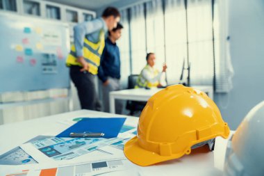 Concept of engineering equipments of protective helmet for competent engineer on piles of paper works. Blurred image of engineers and employer working together at the desk.