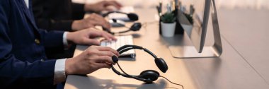 Panorama focus hand holding headset on call center workspace desk with blur background of operator team or telesales representative engaging in providing client with customer support service. Prodigy clipart