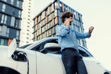 Progressive businessman talking on the phone, leaning on electric car recharging with public EV charging station, apartment condo residential building on the background as green city lifestyle.