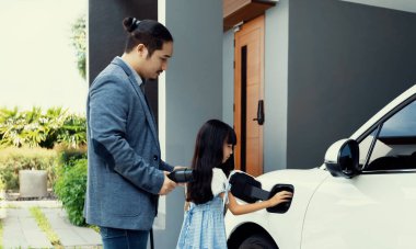 Progressive father and daughter plugs EV charger from home charging station to electric vehicle. Future eco-friendly car with EV cars powered by renewable source of clean energy.