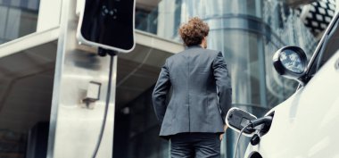 Below view of progressive businessman with electric car recharging at public charging station at modern city residential building. Eco friendly rechargeable EV car powered by alternative clean energy.