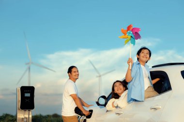 İlerici mutlu aile kavramı elektrikli araçla rüzgar çiftliğinde eğleniyor. Rüzgar türbini jeneratöründen gelen temiz yenilenebilir enerji ile şarj edilen elektrikli araç..