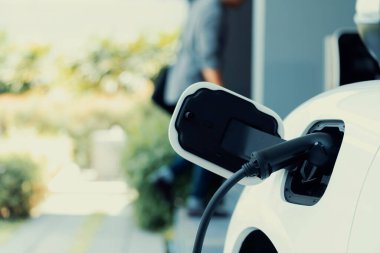 Focus electric car charging at home charging station with blurred progressive man walking in the background. Electric car using renewable clean for eco-friendly concept.