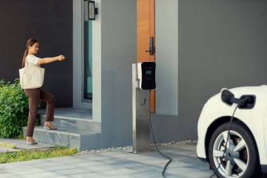 Asyalı bir kadın garajında uzaktan kumandalı araba anahtarıyla elektrikli arabayı kilitliyor, EV şarj istasyonunda şarj pili var. Alternatif temiz enerji günlük hayatta yenilikçi yaşam tarzı kavramı olarak uygulanır.