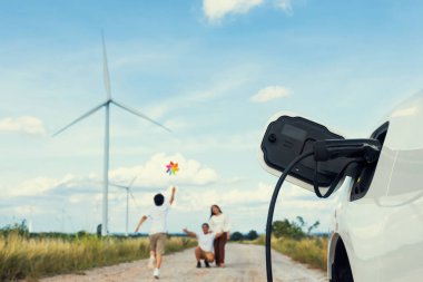 Concept of progressive happy family holding windmill toy and relax at wind farm with electric vehicle. Electric vehicle driven by clean renewable energy from wind turbine generator to charger station.