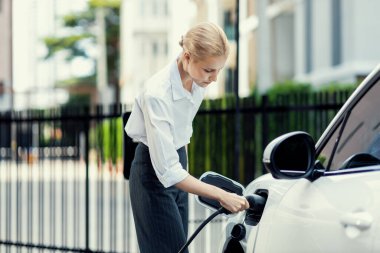 Progressive businesswoman insert charger plug from charging station to her electric vehicle with apartment condo building in background. Eco friendly rechargeable car powered by sustainable energy.