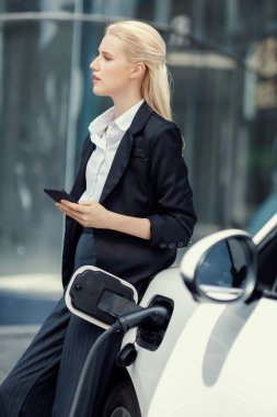 Businesswoman wearing black suit using smartphone, leaning on electric car recharge battery at charging station in city residential building with condos and apartment. Progressive lifestyle concept.