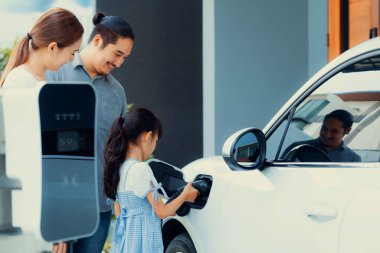 Progressive young parents and daughter with electric vehicle and home charging station. Green and clean energy from electric vehicles for healthy environment. Eco power from renewable source at home.