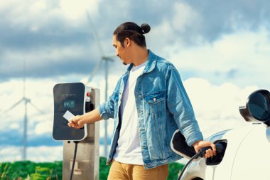 Elektrikli arabasıyla ilerici bir adam, EV arabası gelecekteki sürdürülebilir enerji kavramı olarak rüzgar türbiniyle yeşil alandaki enerji şarj etme istasyonundan enerji yüklüyor. Enerji üreteci olan elektrikli bir araç..