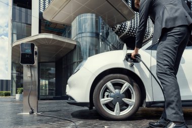 Below view closeup image of progressive black suit businessman recharge battery of his electric vehicle from public charging station. Renewable and alternative energy powered car concept.