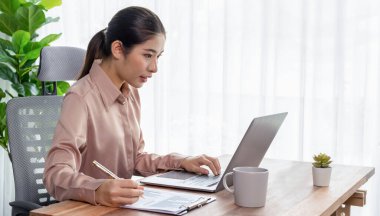Modern ofis masasındaki Asyalı hevesli genç iş kadını çalışmak ve notlar yazmak için dizüstü bilgisayar kullanıyor. Çalışkan ve çekici ofis hanımı ofisinde bilgisayar defteri üzerinde çalışıyor..