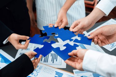 Closeup top view business team of office worker putting jigsaw puzzle together over table filled with financial report paper in workplace with manager to promote harmony concept in meeting room.
