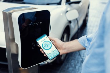Closeup progressive businessman look at EV cars battery status application on smart phone screen at public parking charging station with power cable plug and renewable energy-powered electric vehicle