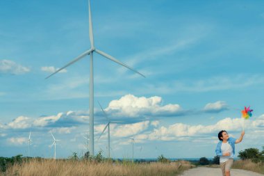 Progressive young asian boy playing with wind pinwheel toy in the wind turbine farm, green field over the hill. Green energy from renewable electric wind generator. Windmill in the countryside concept