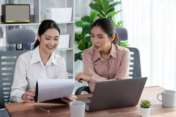 Two young office lady colleagues collaborating in modern office workspace, engaging in discussion and working together on laptop, showcasing their professionalism as modern office worker. Enthusiastic