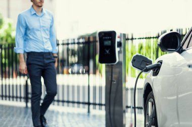 Progressive eco-friendly concept of focus parking EV car at public electric-powered charging station in city center with blur businessman walking in the background.