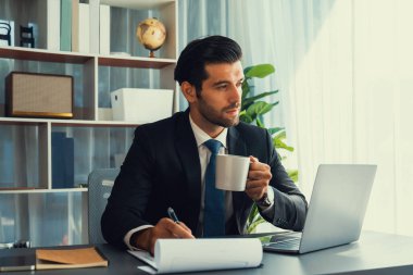 Modern ofis masasındaki modern profesyonel iş adamı elinde kahveyle çalışmak için dizüstü bilgisayar kullanıyor. Ofisinde bilgisayar defteri üzerinde çalışan çalışkan bir ofis çalışanı. coşkulu