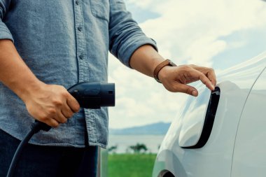 Progressive natural scenic where hand insert charging plug to electric vehicle from charging station with natural background. Future energy concept of EV car powered by sustainable electric energy.