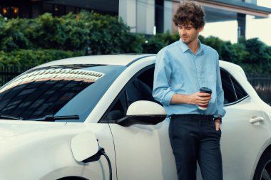 Progressive eco-friendly concept of parking EV car at public electric-powered charging station in city with blur background of businessman leaning on recharging-electric vehicle with coffee.