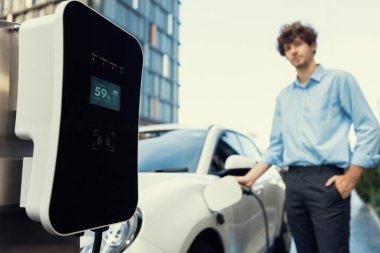 Focus public EV charging station in city residential area with blur background of businessman charging electric vehicle. Progressive lifestyle of ecological concern of city-dwelling individual.