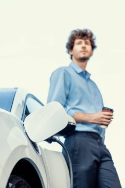 Progressive eco-friendly concept of focus parking EV car at public electric-powered charging station in city with blur background of businessman leaning on recharging-electric vehicle with coffee.